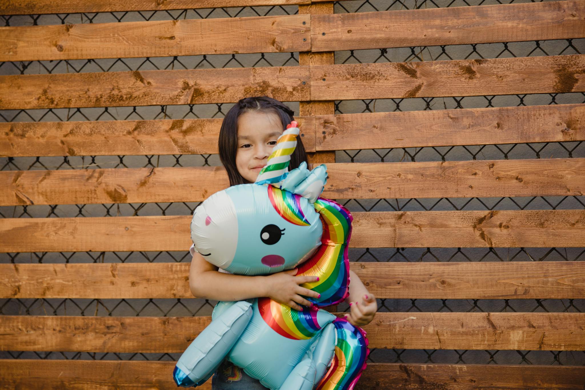 A charming scene of a child holding a colorful unicorn balloon against a wooden fence backdrop.